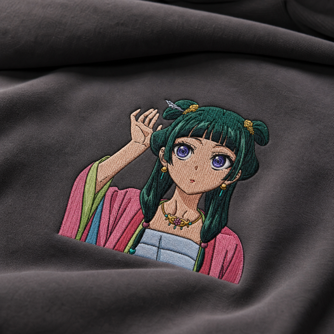 Real-photo premium angled folded-fabric detail of Maomao embroidery on dark gray hoodie fabric.